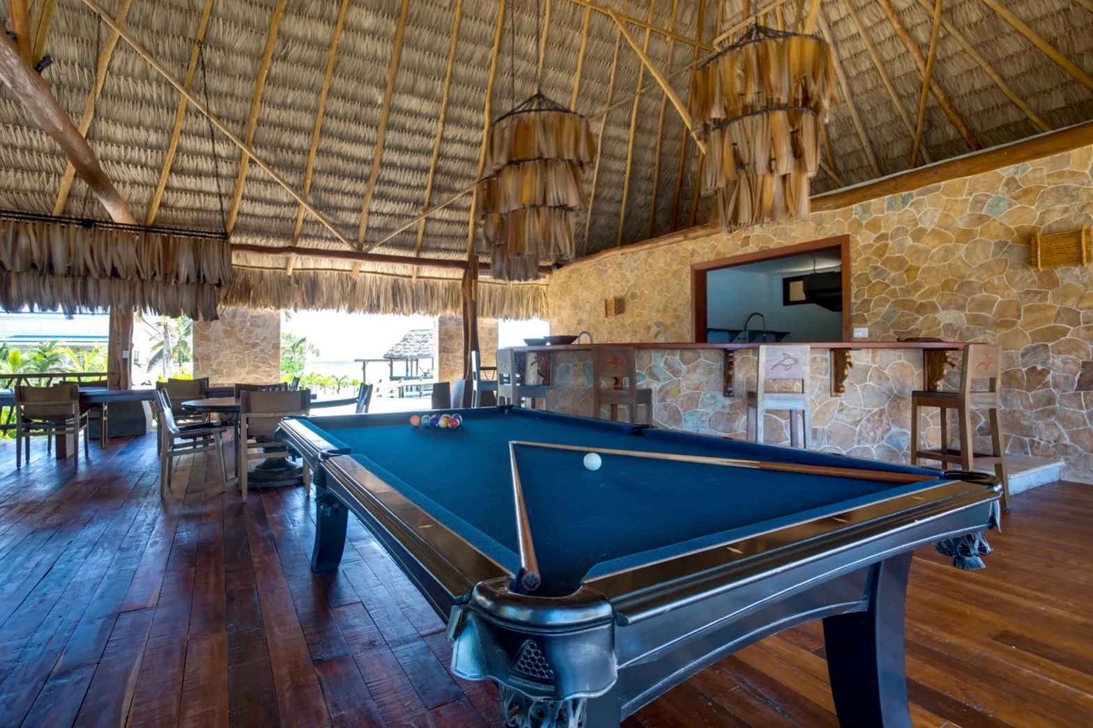 A pool table fills an open-sided recreation room beneath a thatched roof.
