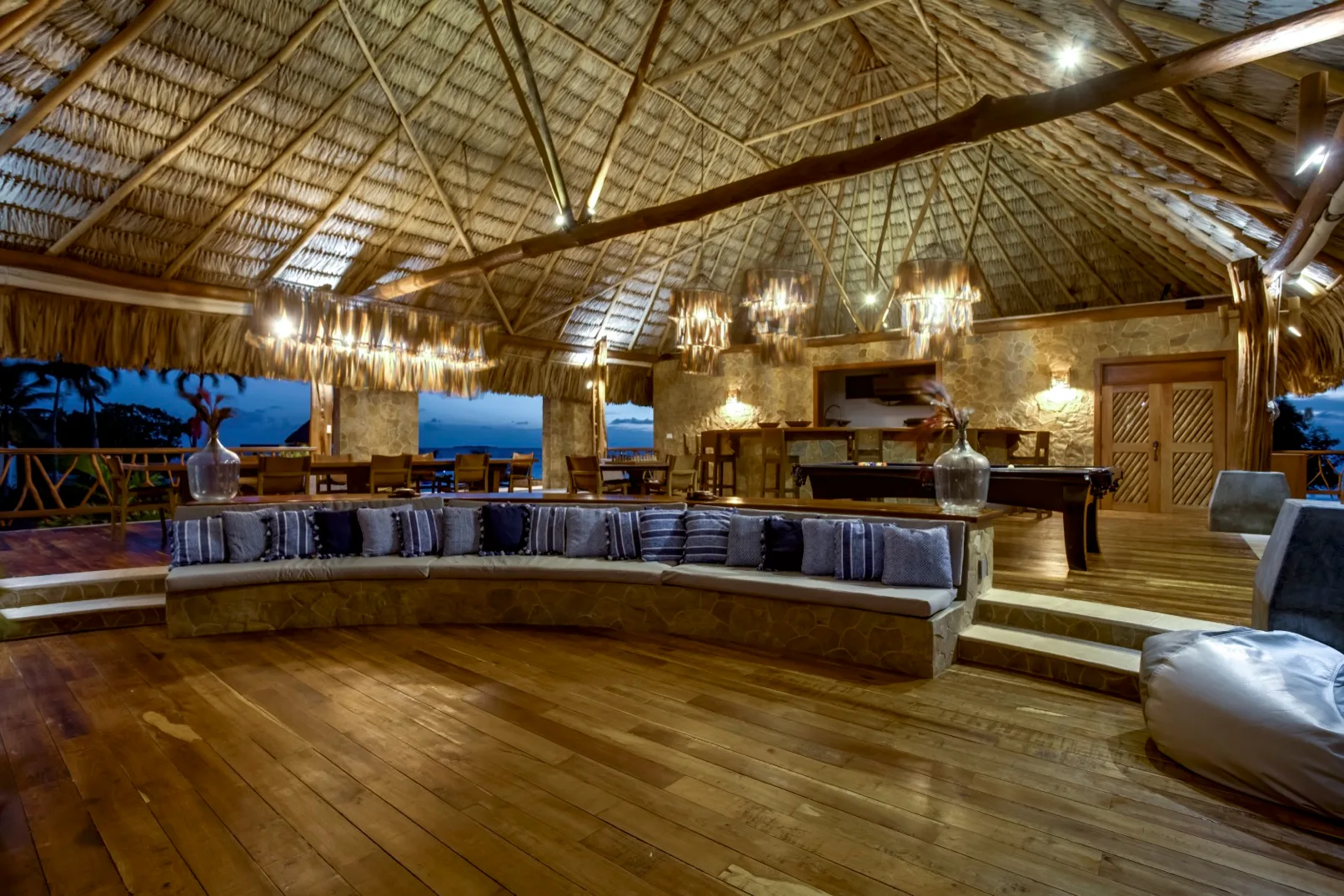 An open-air lounge with a thatched roof, wooden floor, and long seating area.