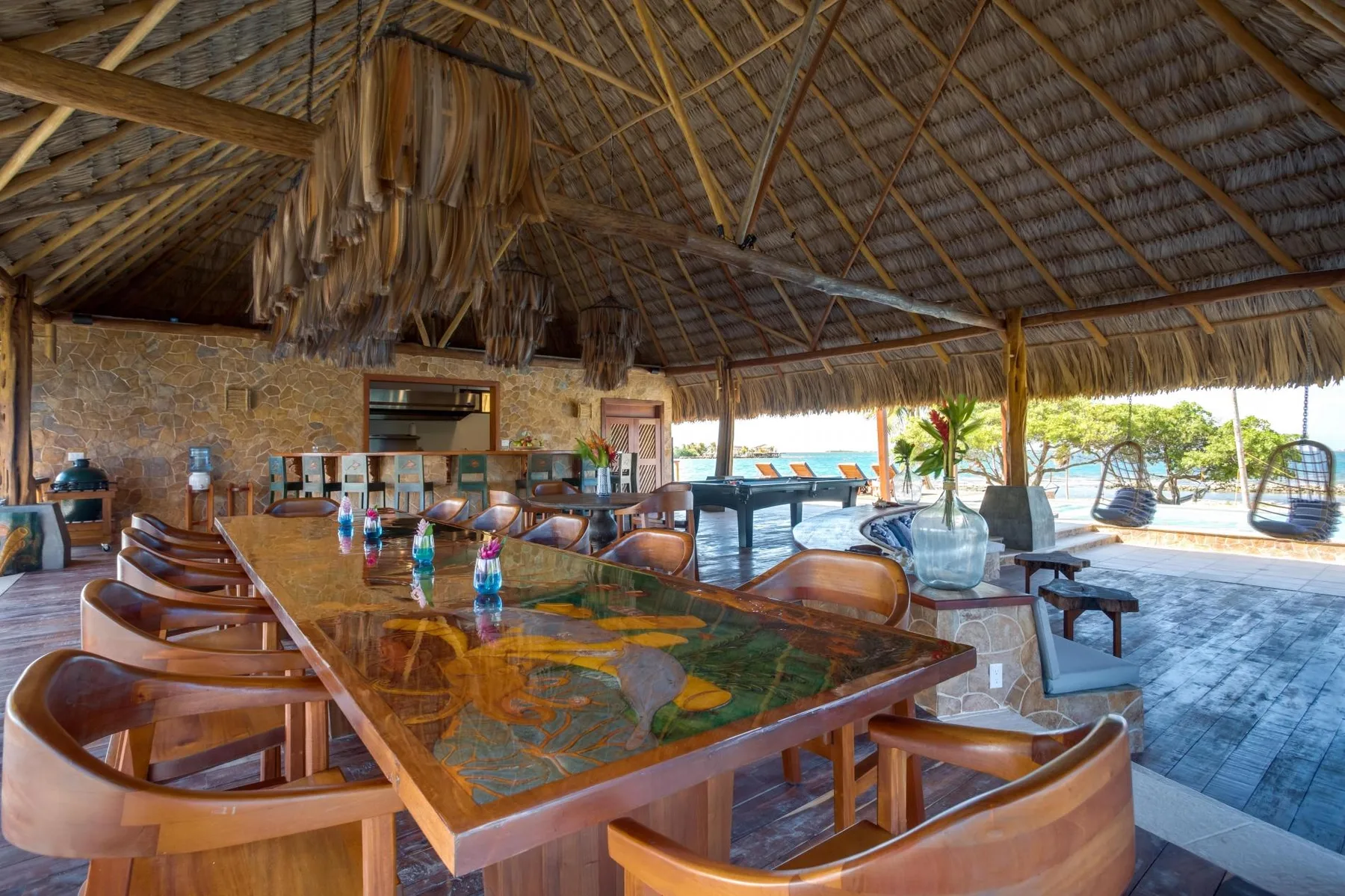An open-air dining area has a long table beneath a thatched roof.