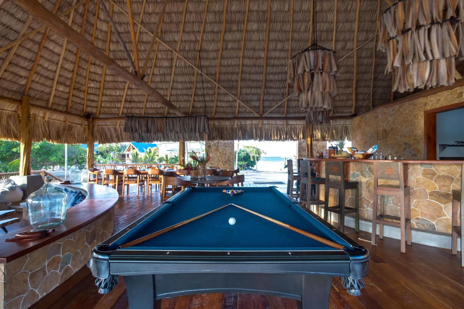 A thatched-roof lounge features a pool table, dining tables, and a stone bar.