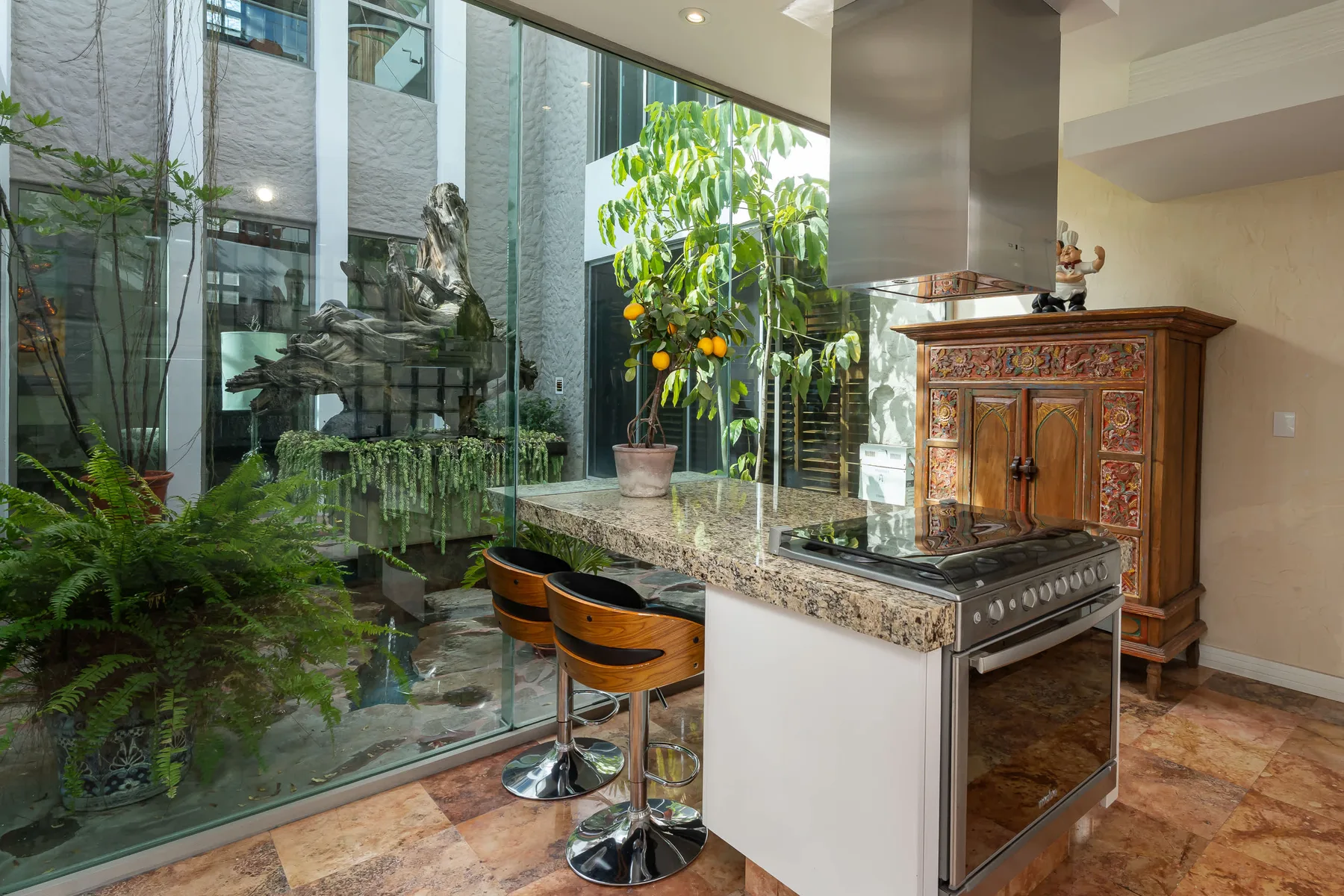 A kitchen island with a cooktop and seating overlooks a glass-walled courtyard garden.