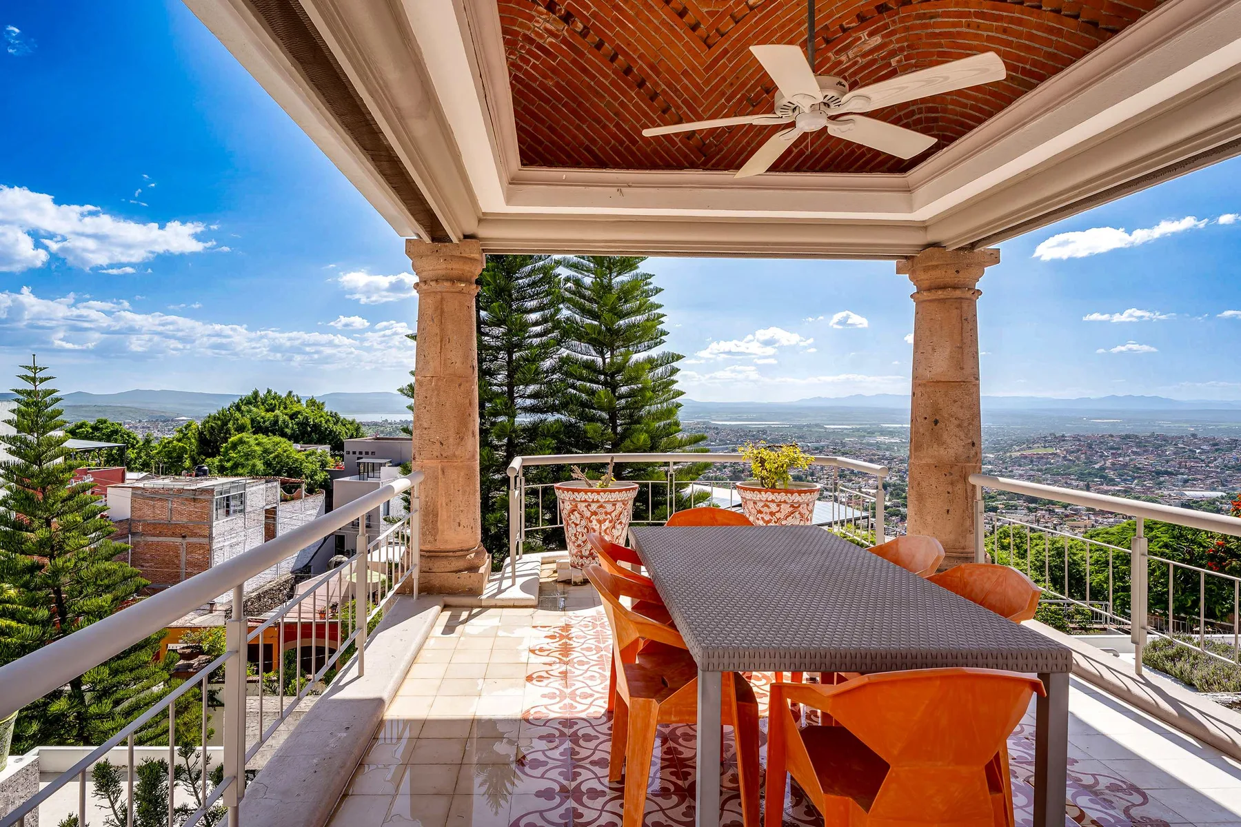 A covered outdoor terrace has a dining table and chairs overlooking the surrounding city.