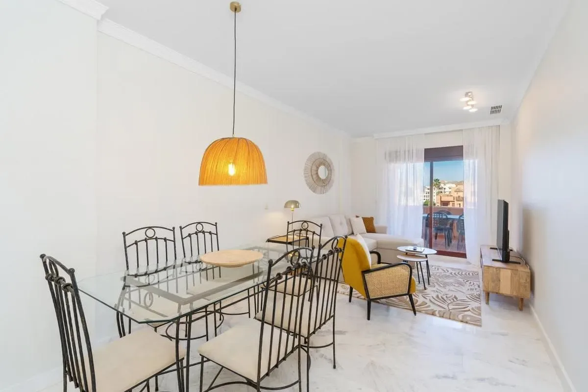 A bright living and dining area with a glass dining table and seating by the window.