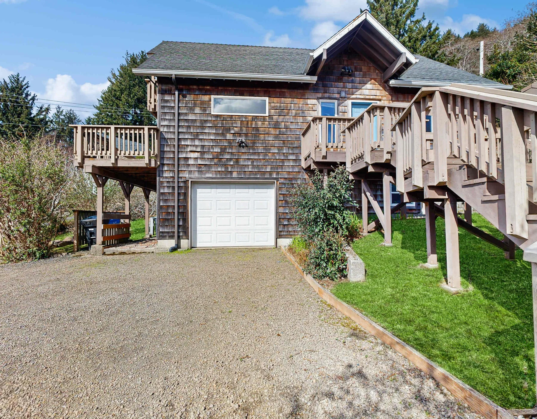 Exterior view of a shingled house with a raised wooden deck and gravel driveway.