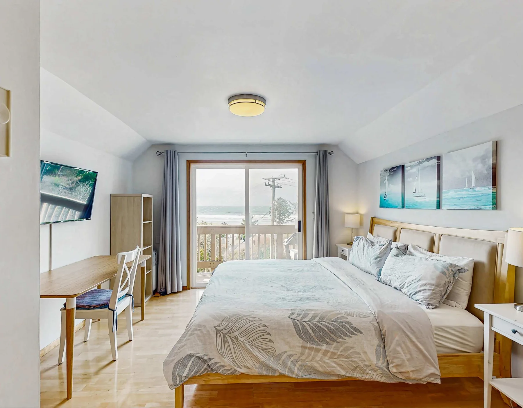 A bright bedroom with a large bed, a desk, and sliding glass doors opening to a balcony.