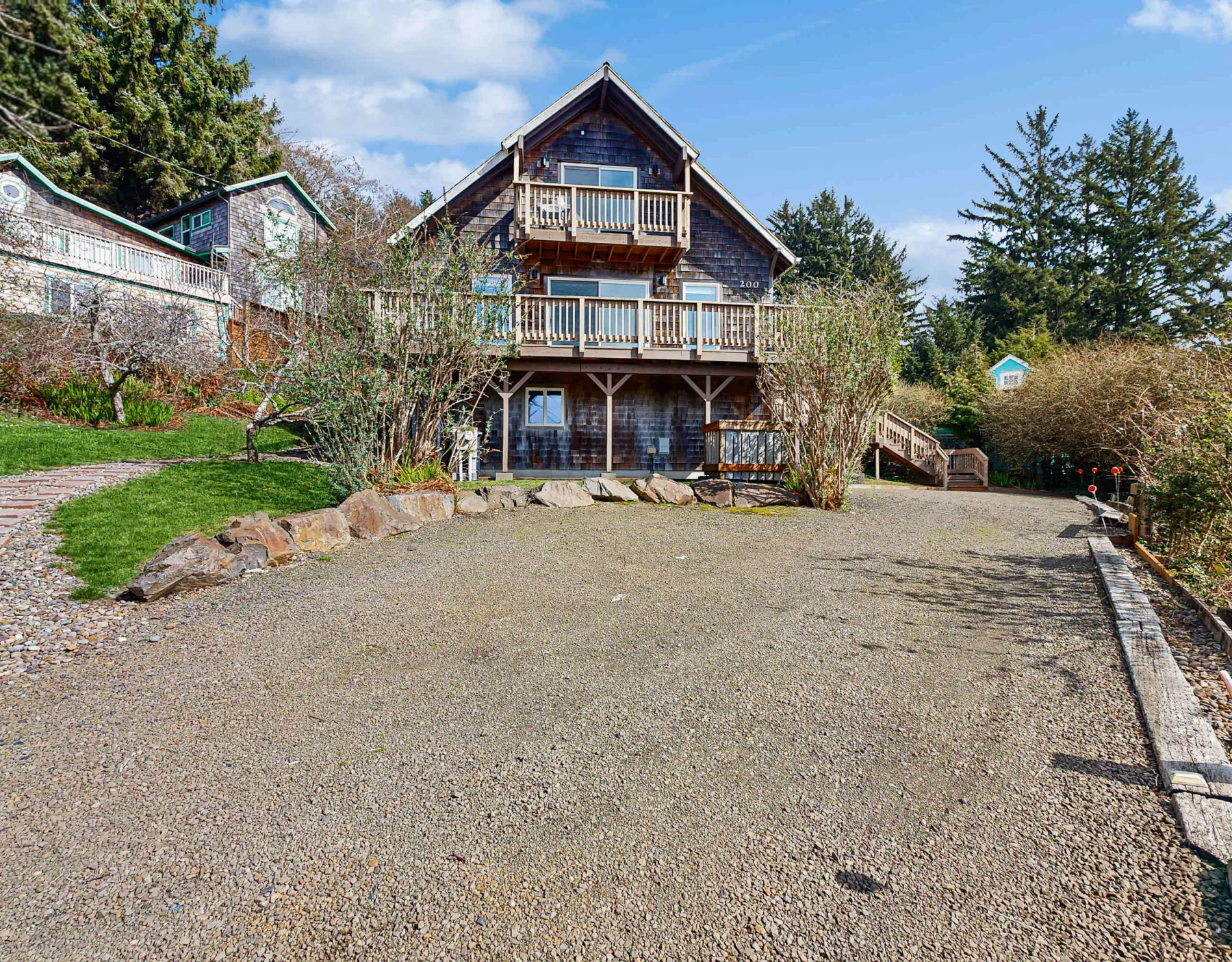A chalet-style house faces a broad gravel courtyard with trees and shrubs.