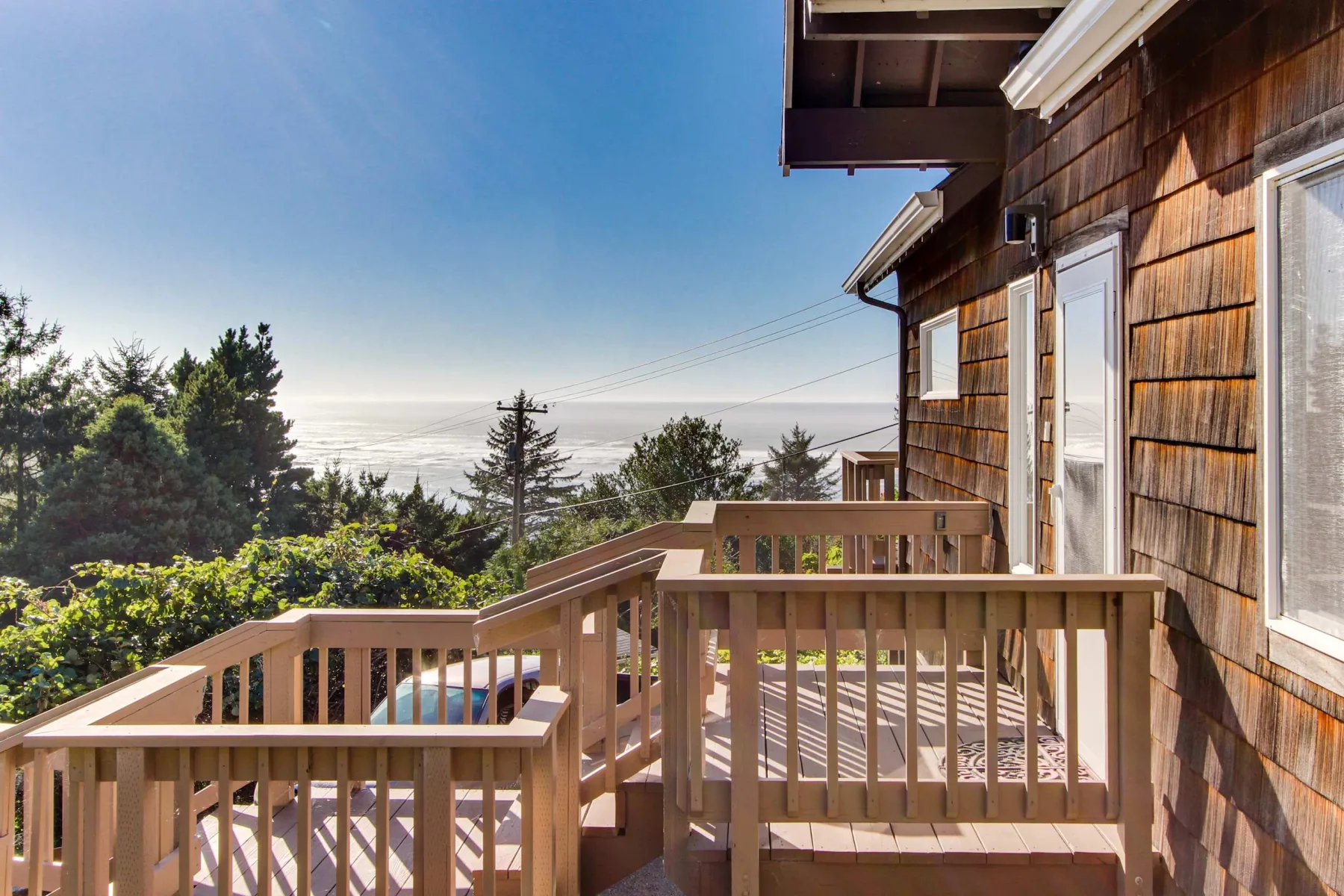 A wooden deck beside a shingled house overlooks trees and a distant ocean.