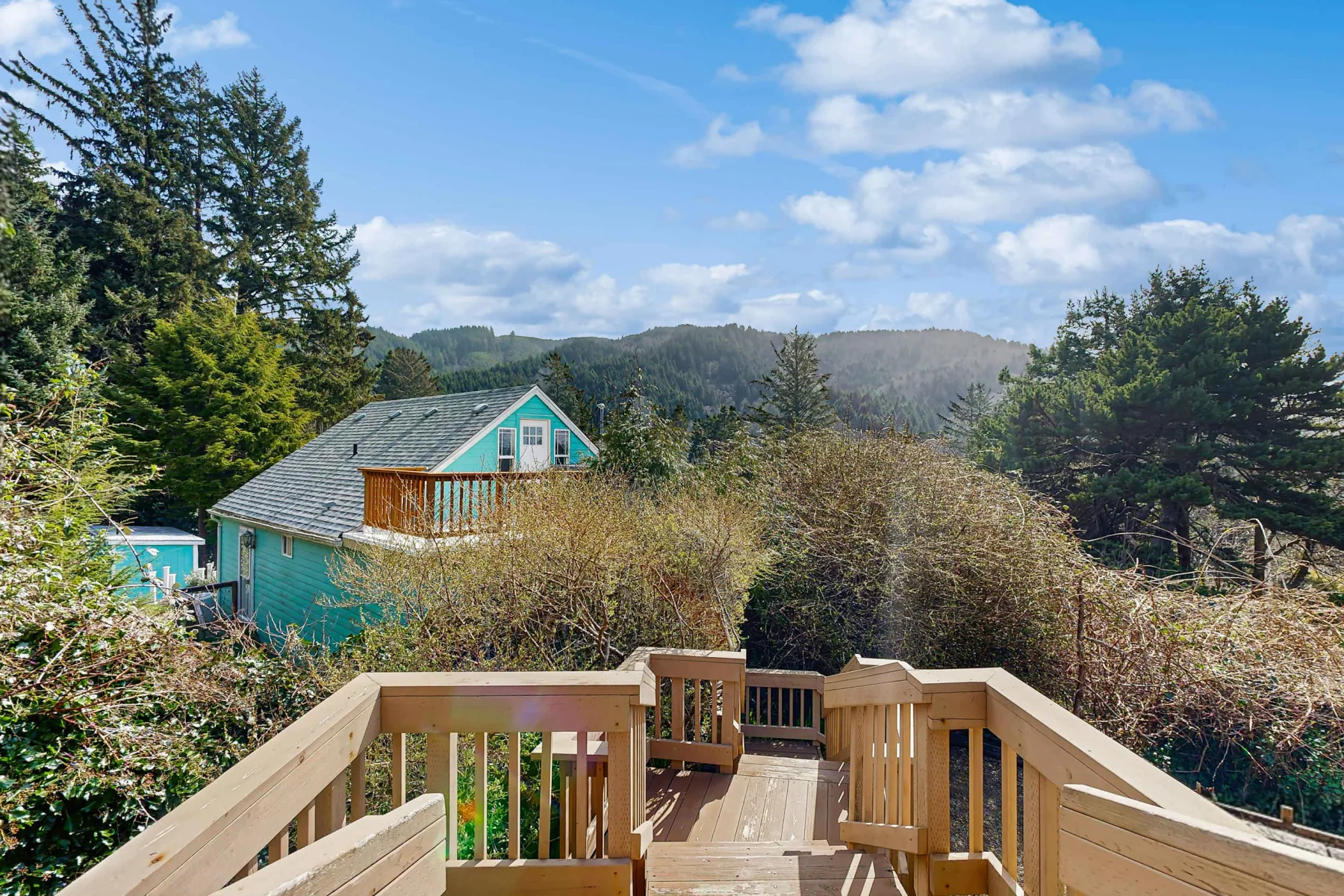 A wooden deck overlooks a teal house, trees, and distant hills beneath a partly cloudy sky.