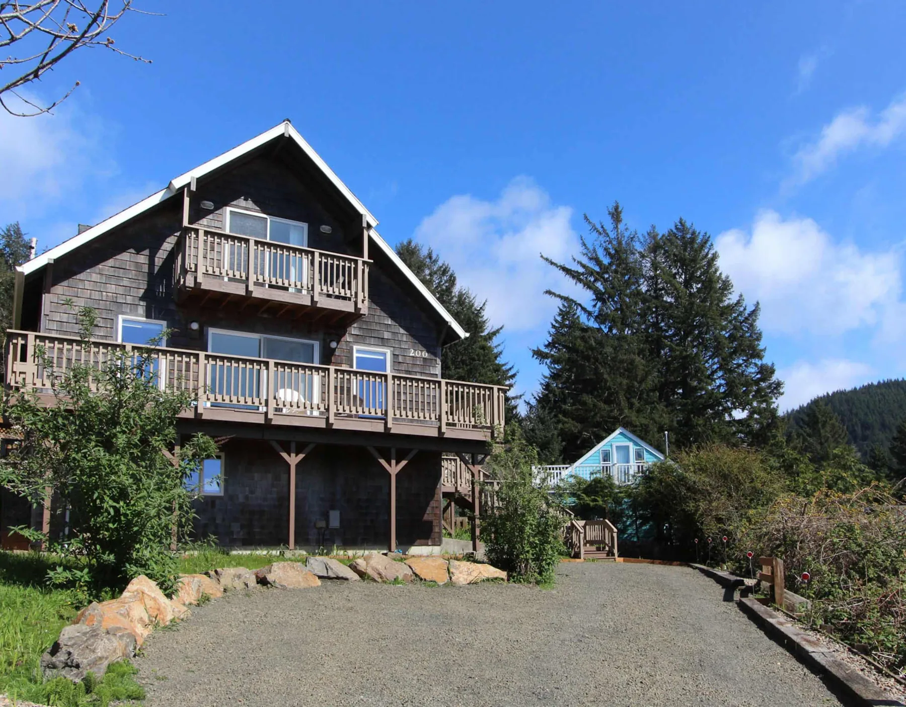 A dark wooden house with two balconies stands beside a gravel driveway.