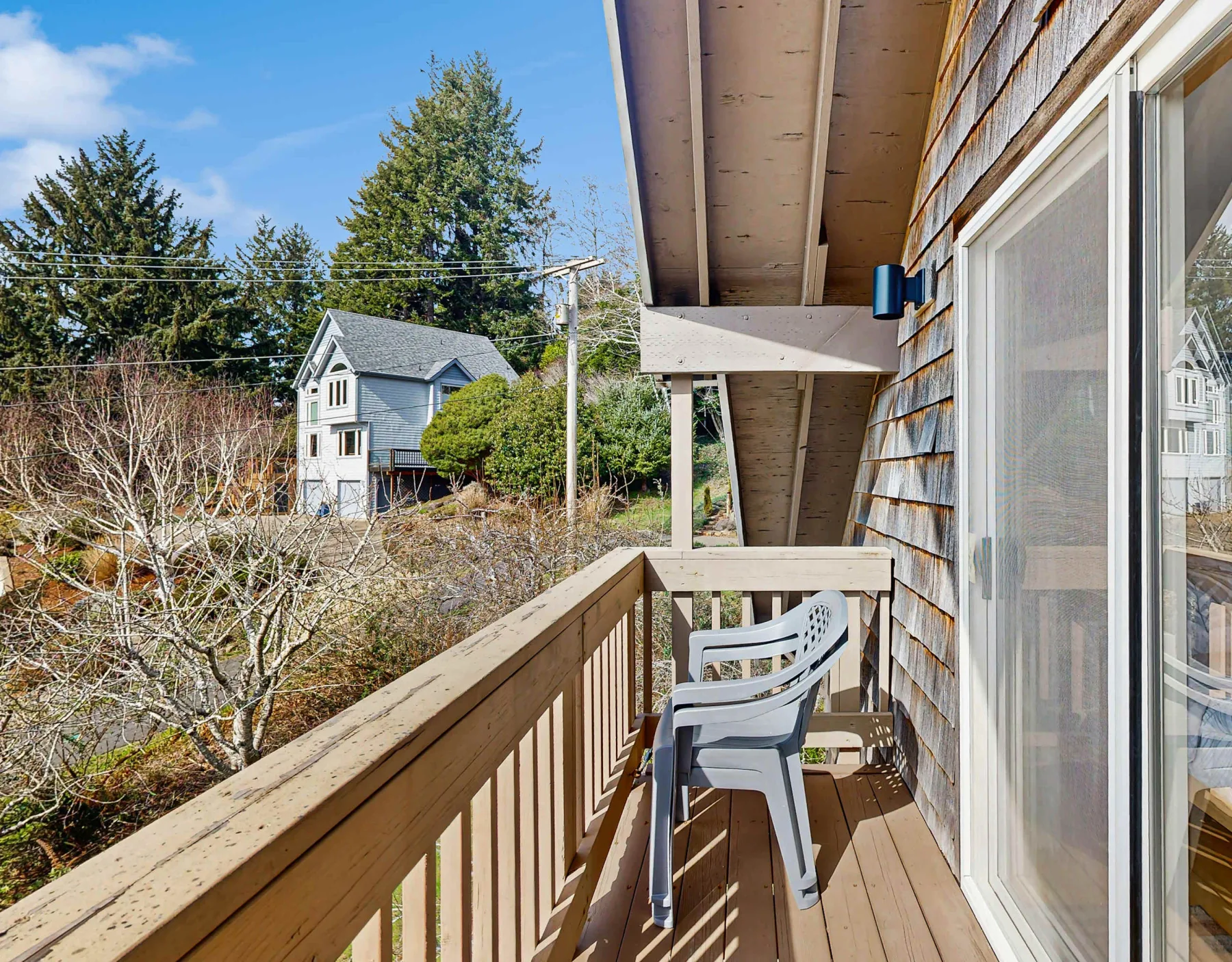 A narrow wooden balcony with seating overlooks trees and a neighboring house.