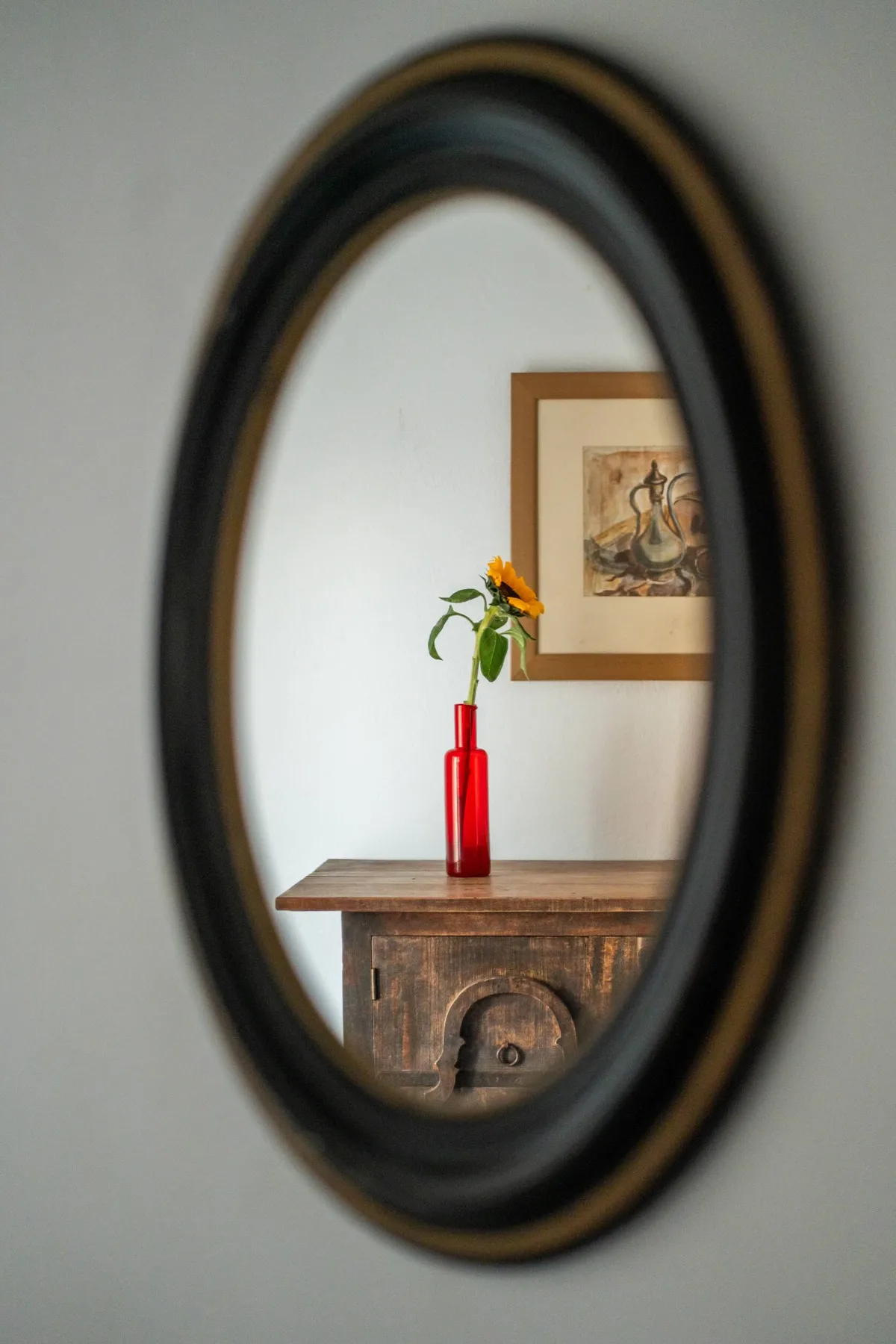A framed artwork and flower in a red vase are reflected in an oval mirror.