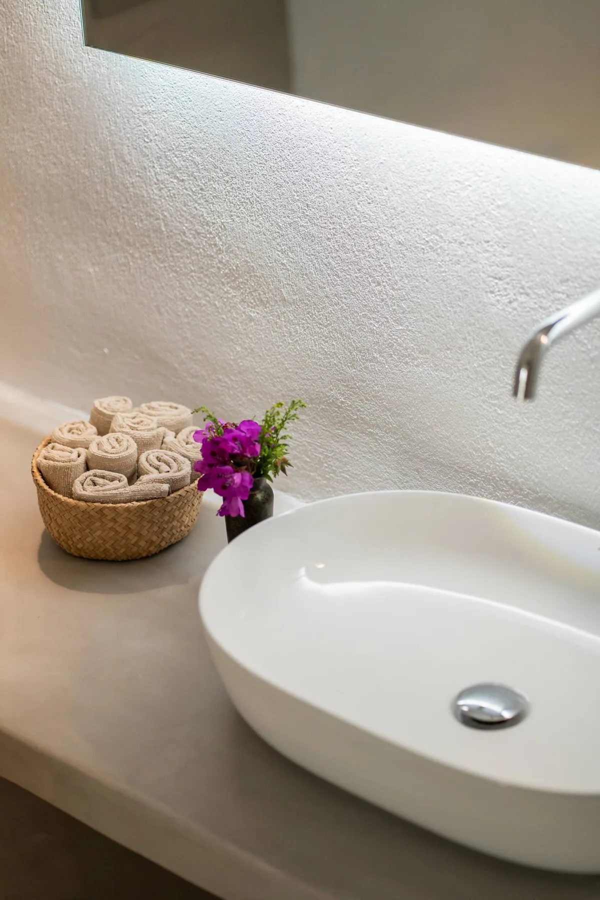 A bathroom vanity with a white basin, rolled towels, and flowers.