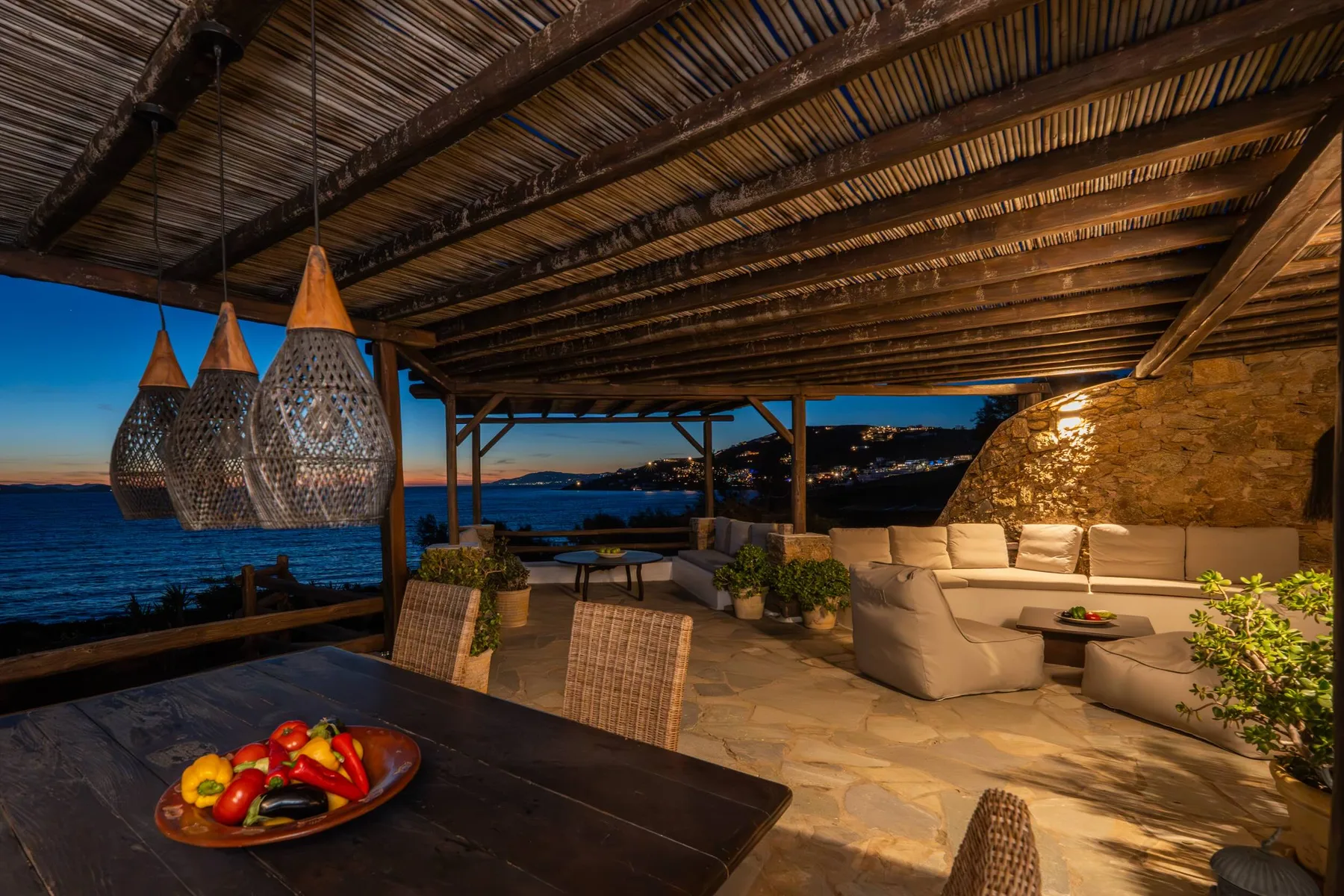 A covered outdoor terrace with lounge seating overlooks the sea at dusk.