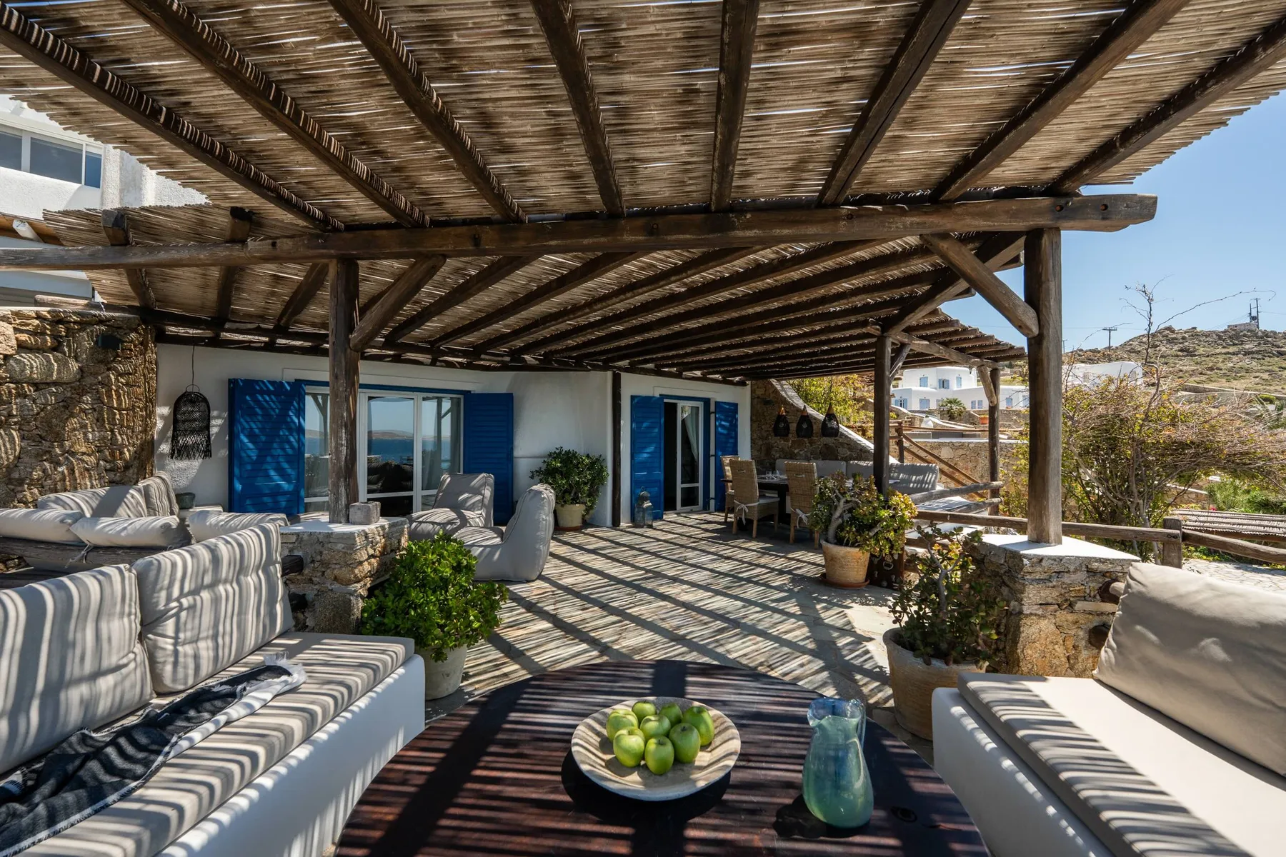 A shaded outdoor patio has cushioned seating, a low table, and a wooden pergola.