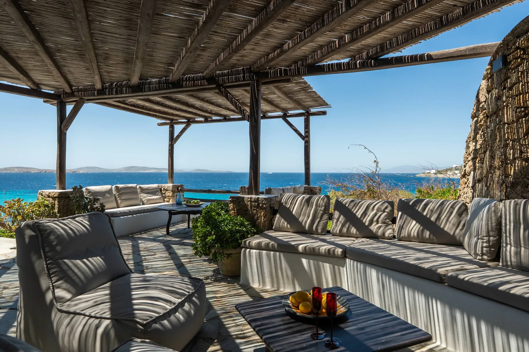 An outdoor lounge under a wooden pergola overlooks the sea.