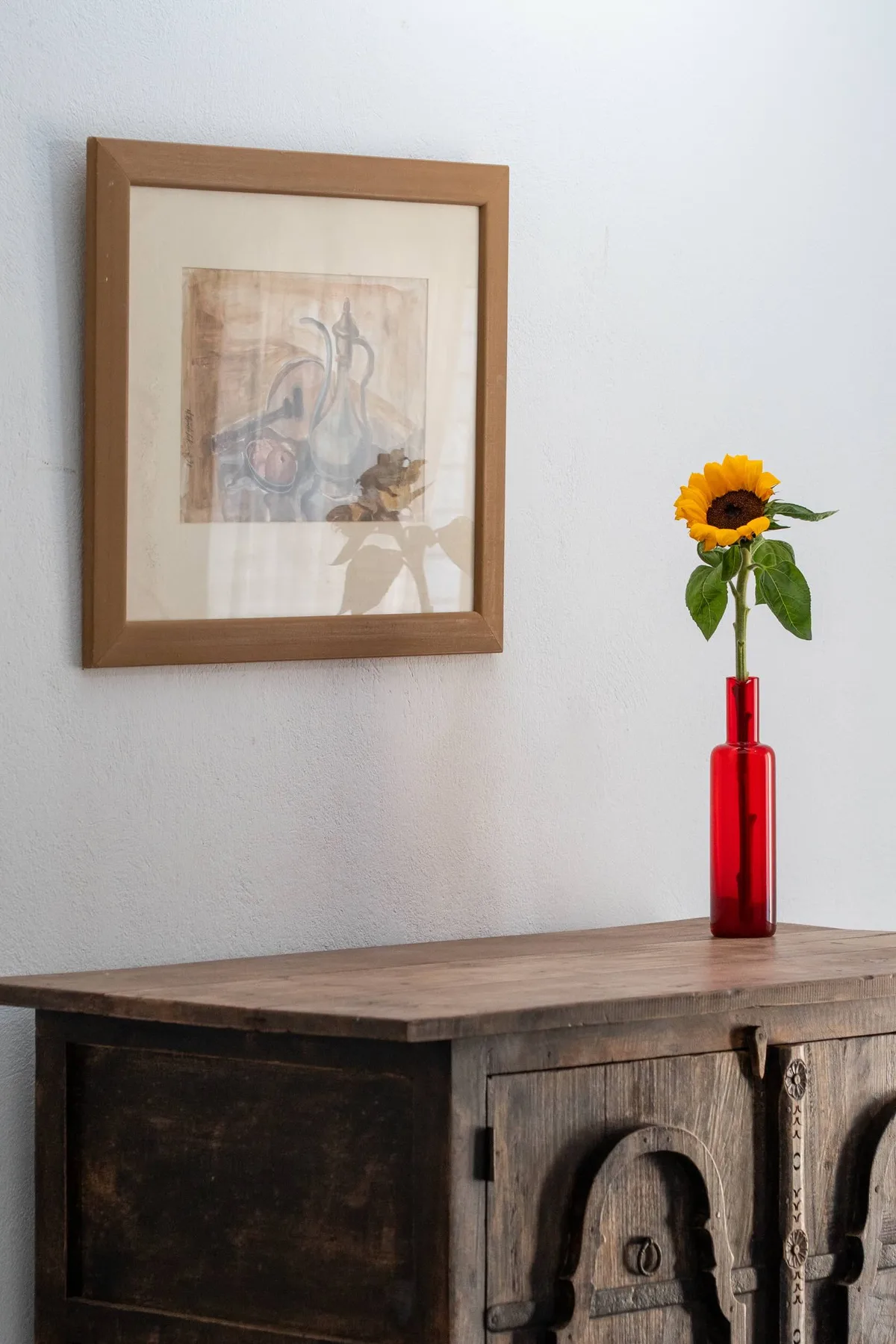A wooden cabinet stands beneath framed artwork, with a sunflower in a red vase.