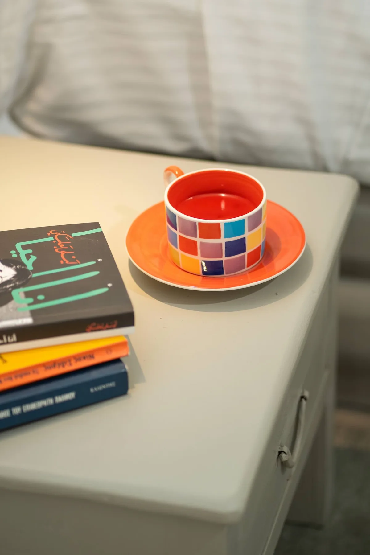 A bedside table holds a colorful cup and saucer beside a stack of books.