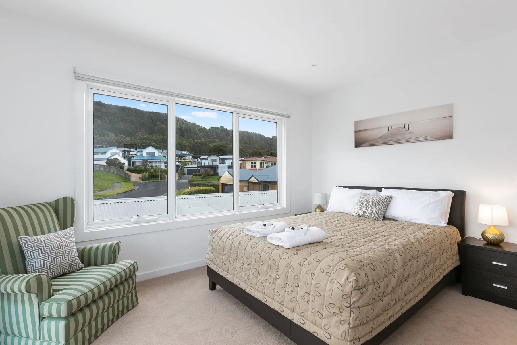 A bedroom with a bed beside a large window overlooking nearby homes.