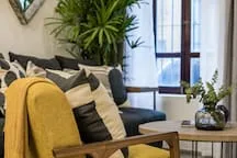 A living room seating area with cushions and plants beside tall windows.