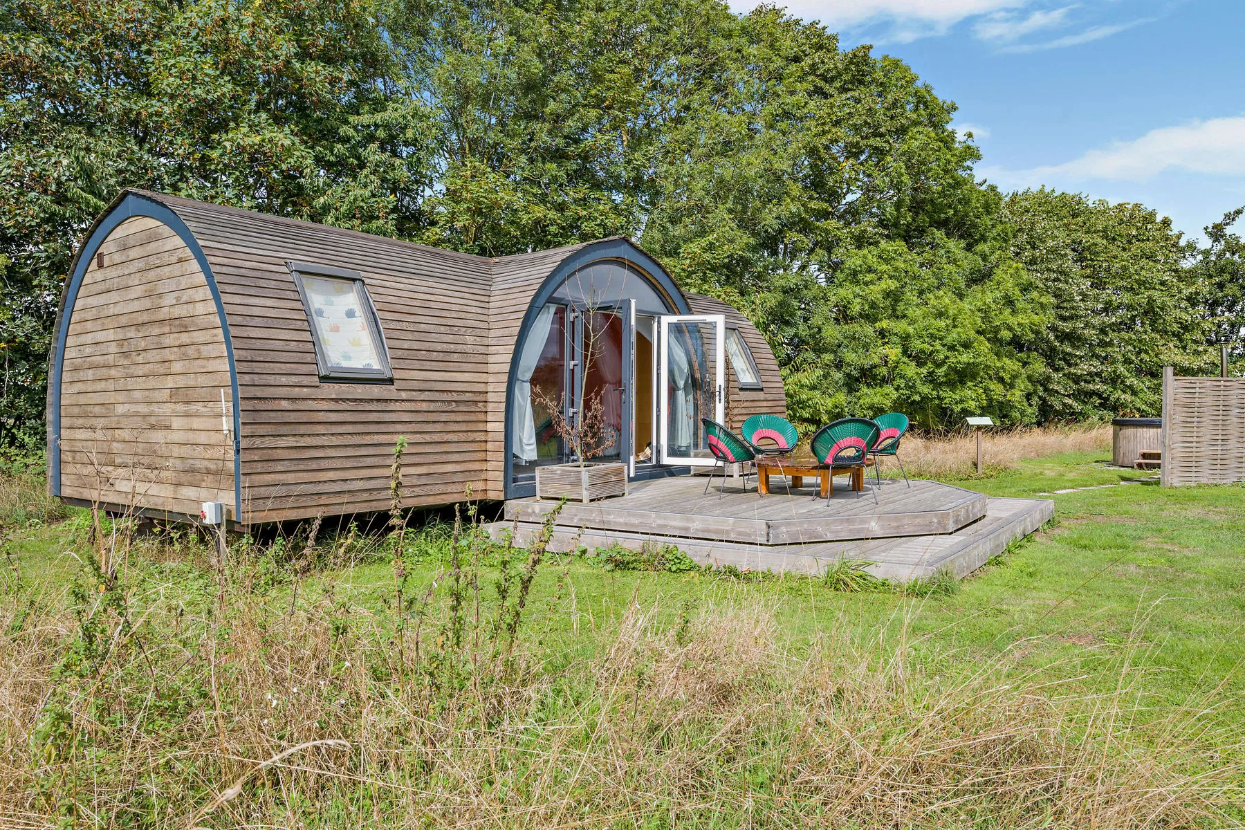 A small curved wooden cabin sits beside a raised deck with colorful outdoor seating.