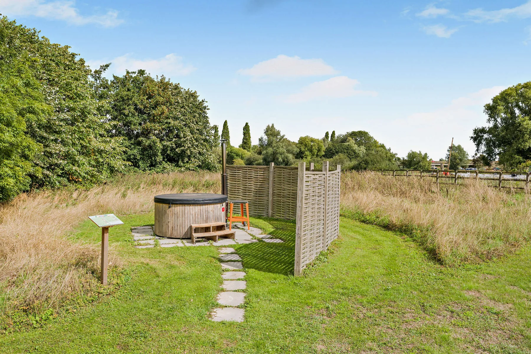 A grassy outdoor area has a wooden hot tub beside a privacy screen and stepping stones.