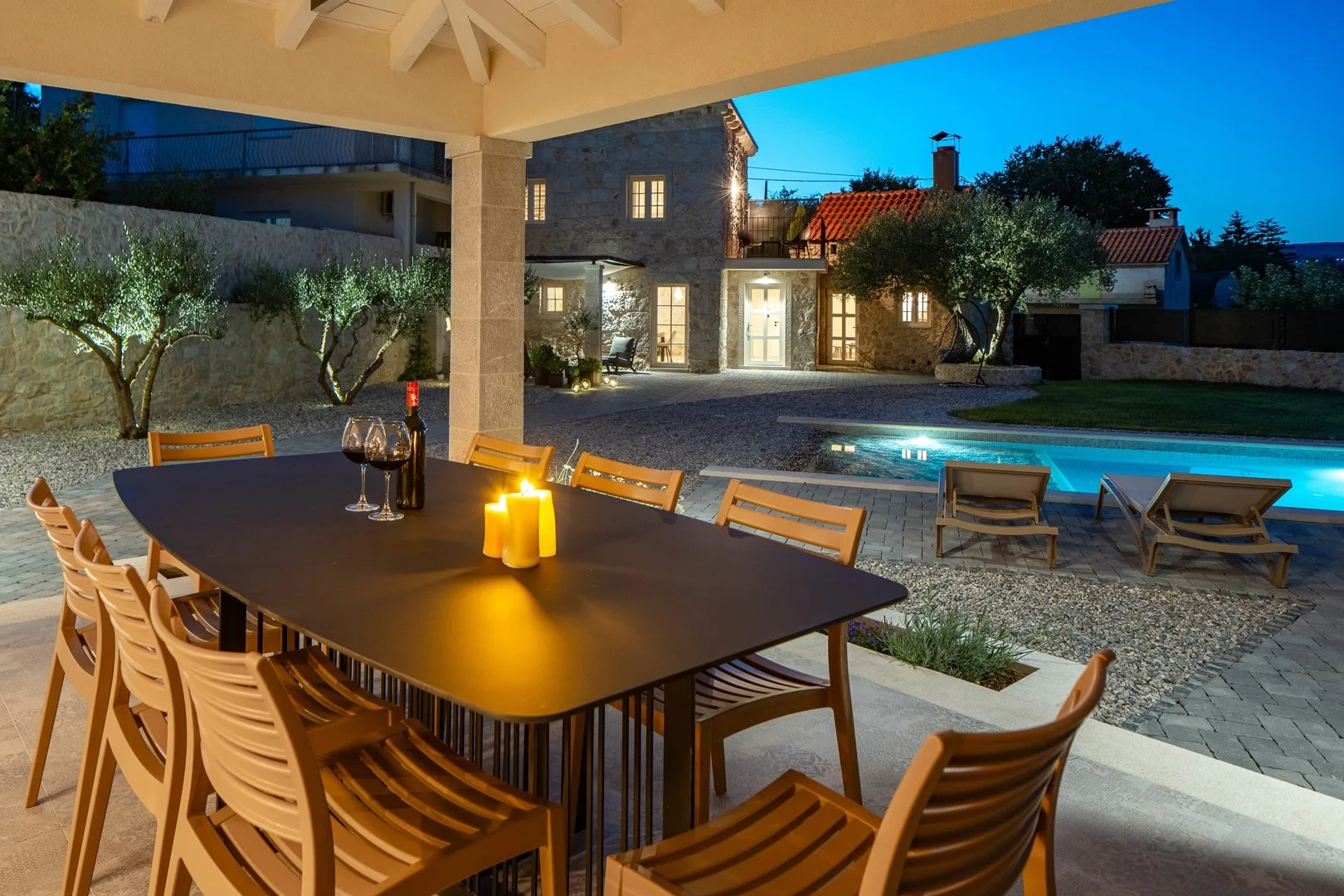 Una zona de comedor exterior cubierta da a una piscina y una casa iluminada al anochecer.