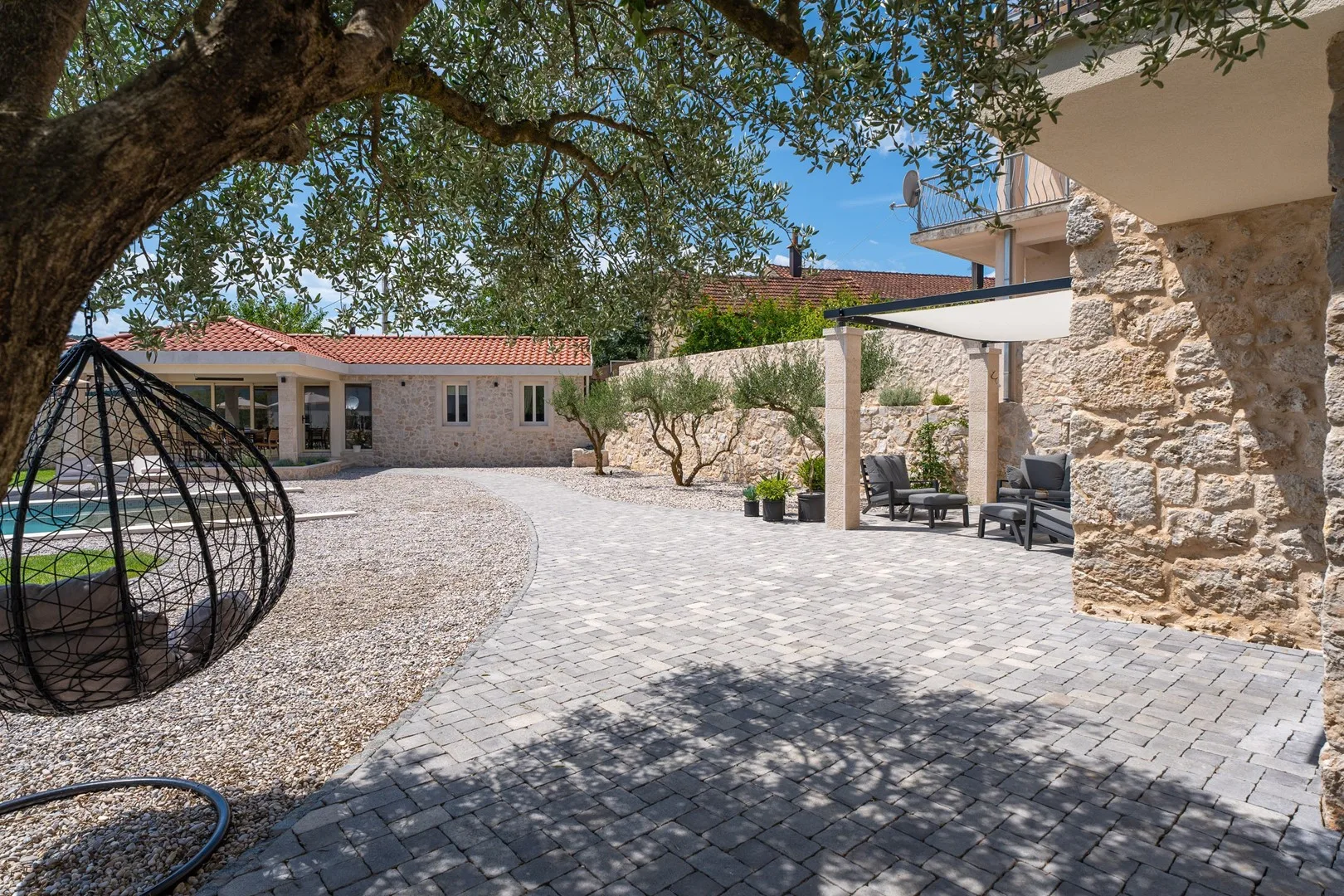 Un patio pavimentado con asientos al aire libre, árboles y una piscina junto a edificios de piedra.