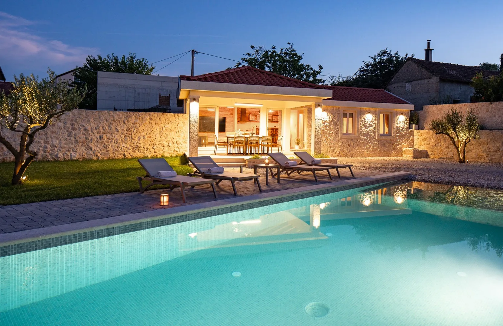 Una piscina y tumbonas están frente a una casa iluminada al anochecer.
