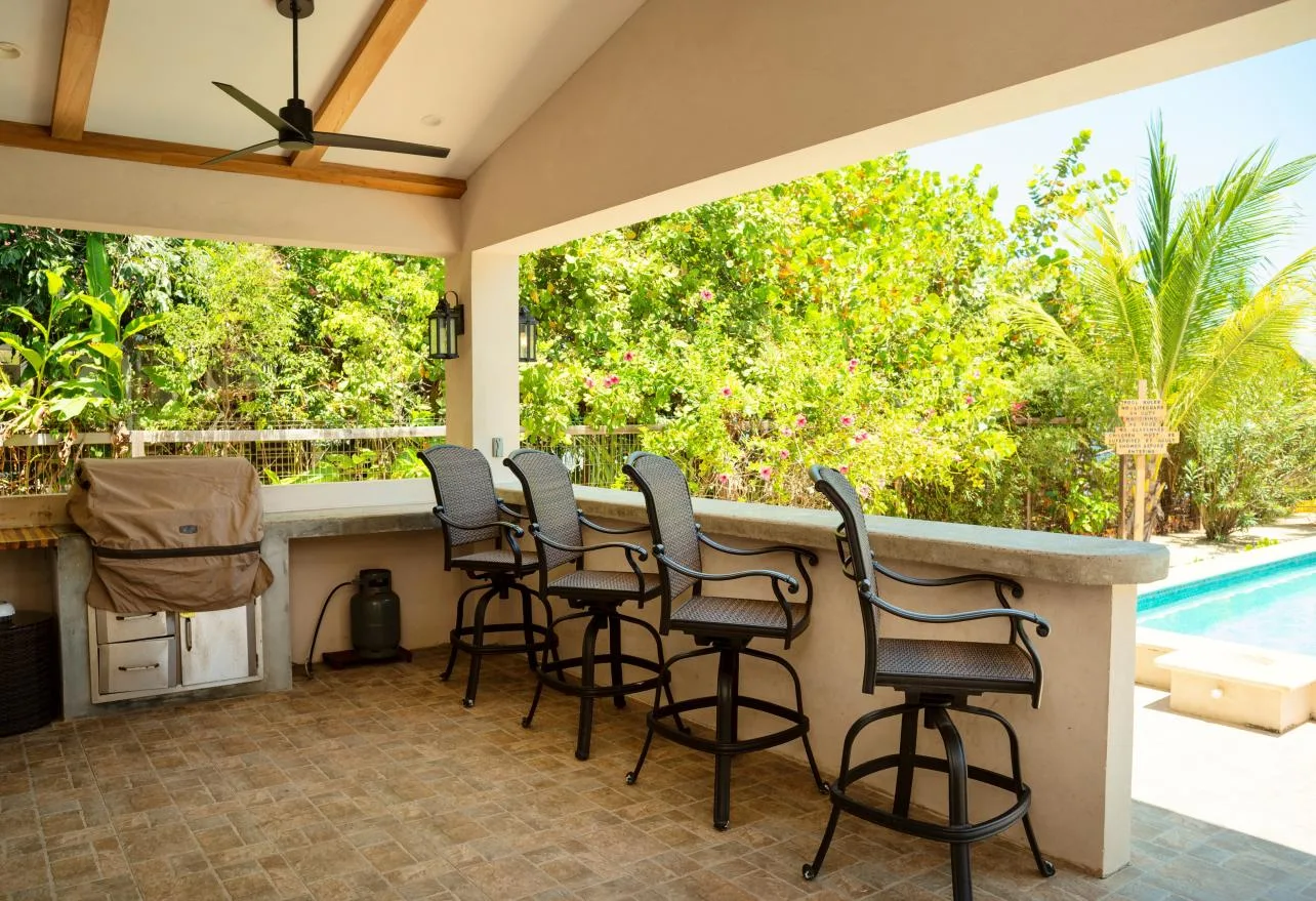 A covered outdoor counter with bar seating overlooks a swimming pool and greenery.