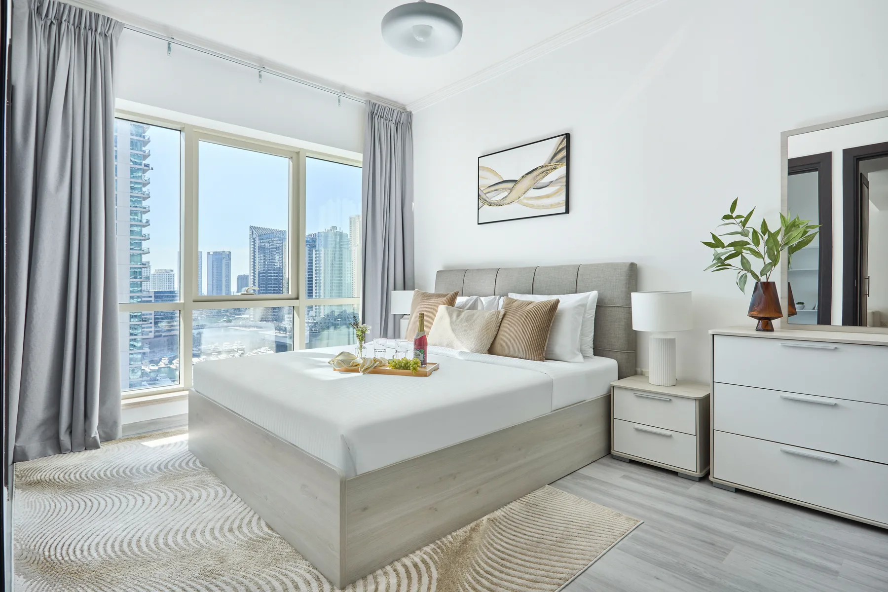 Un dormitorio luminoso con una cama grande junto a amplias ventanas con vistas a edificios de la ciudad.