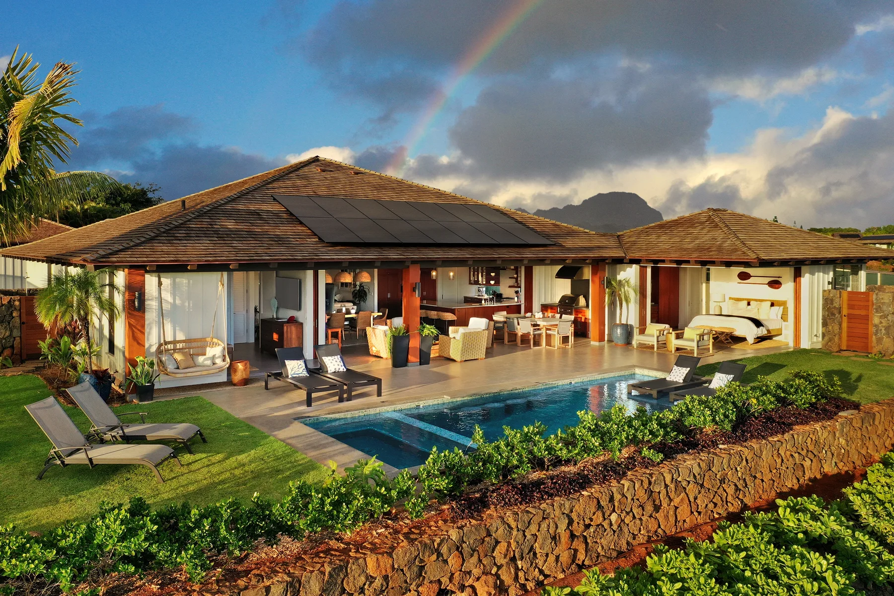 A single-story house opens onto a swimming pool and landscaped garden beneath a rainbow.
