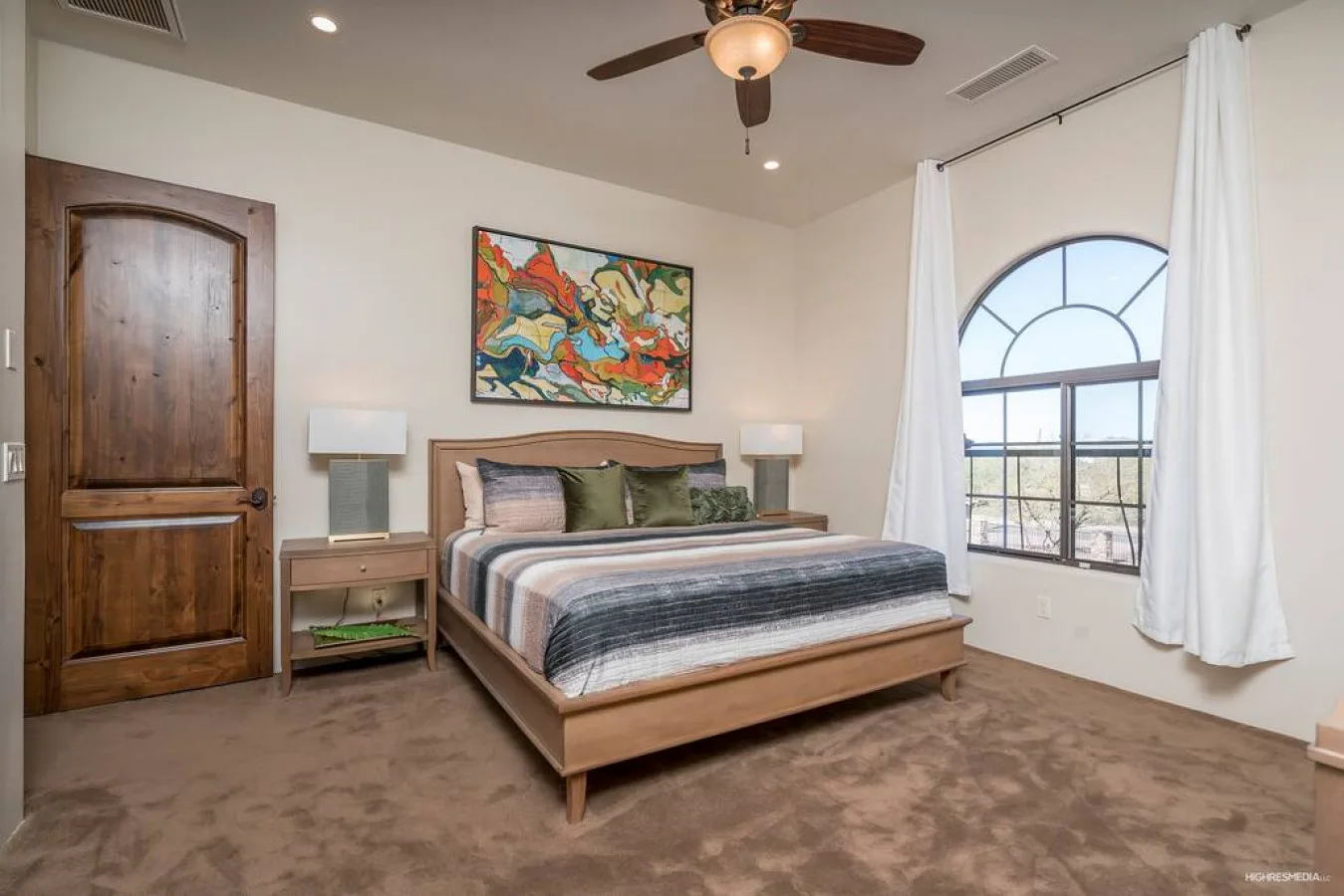 A bedroom with a wooden bed, colorful artwork, and an arched window.