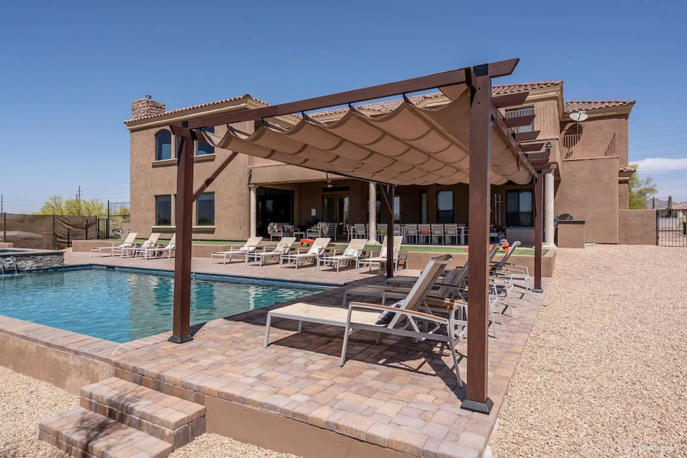 A swimming pool beside a patio with lounge seating and a shaded pergola.