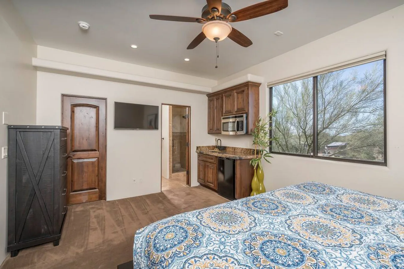 A bedroom with a patterned bedspread, wooden cabinets, and a large window.