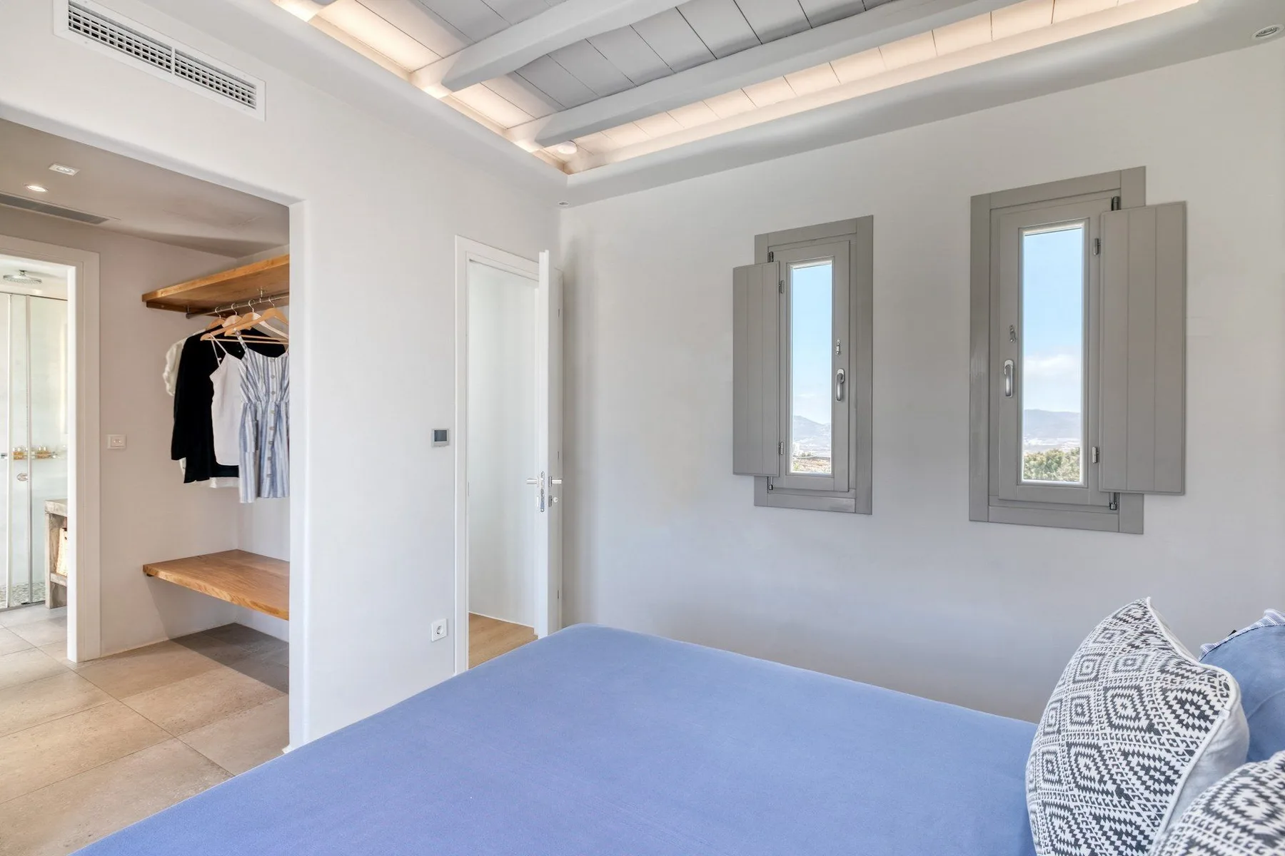 A bedroom with a blue bedspread, two windows, and an open wardrobe.