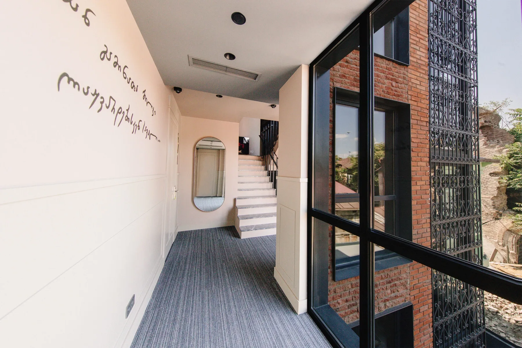 Espacio representativo del hotel: Un pasillo con escaleras pasa junto a grandes ventanas con vistas a un edificio de ladrillo.