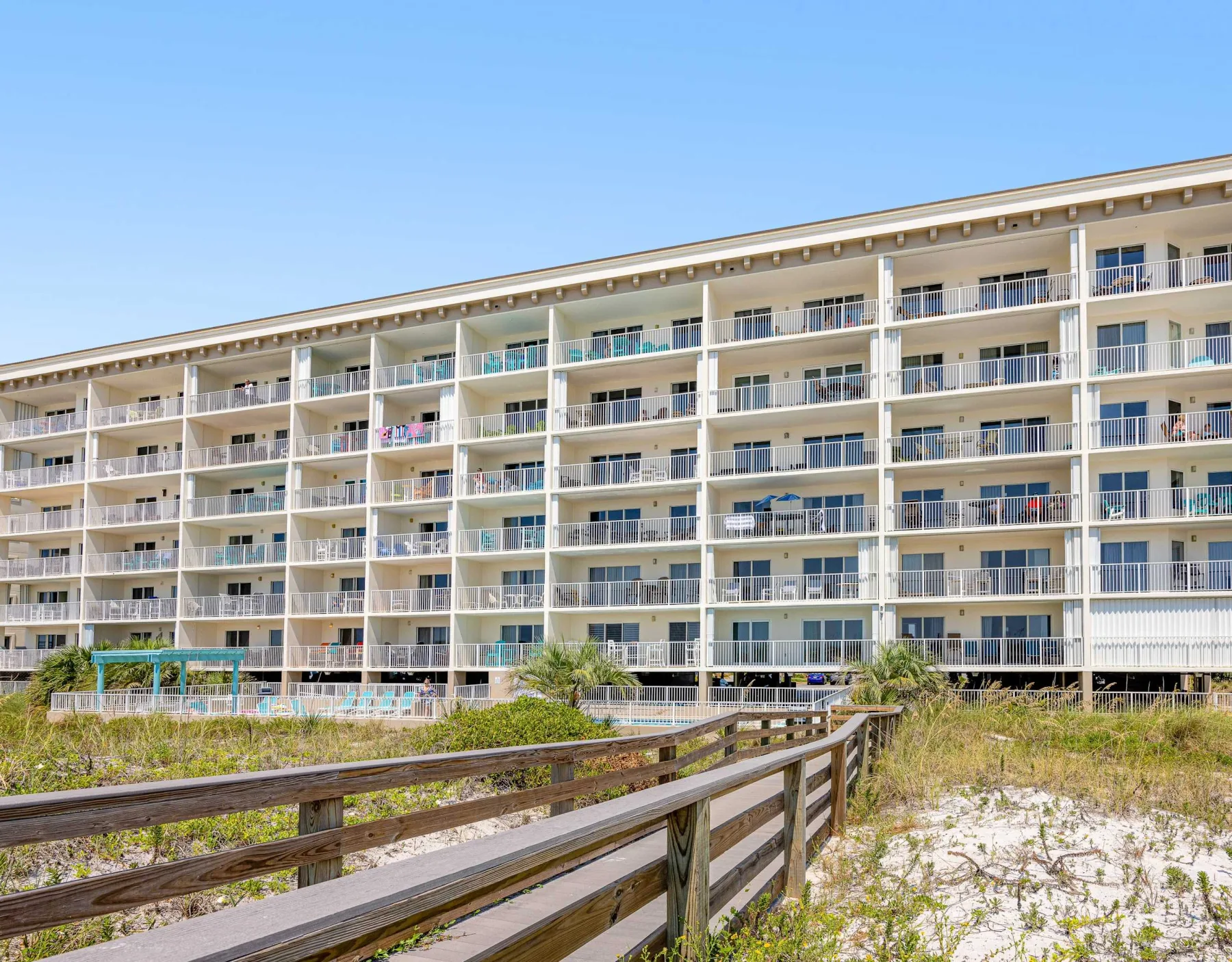Zona común: Un edificio de apartamentos junto a la playa se alza detrás de una pasarela de madera entre vegetación costera.
