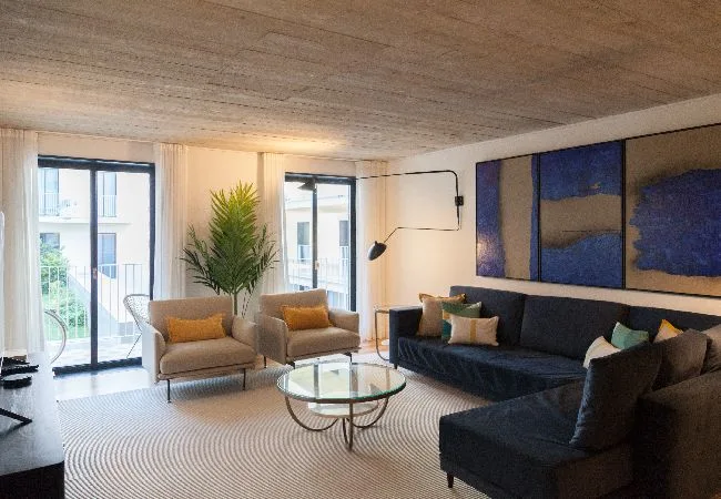 Una sala de estar con asientos oscuros, dos sillones y una mesa de centro de vidrio.