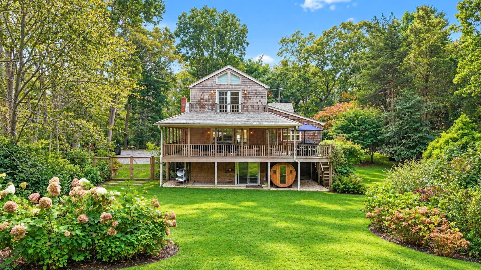 A two-story house with a broad wooden deck sits amid trees and gardens.