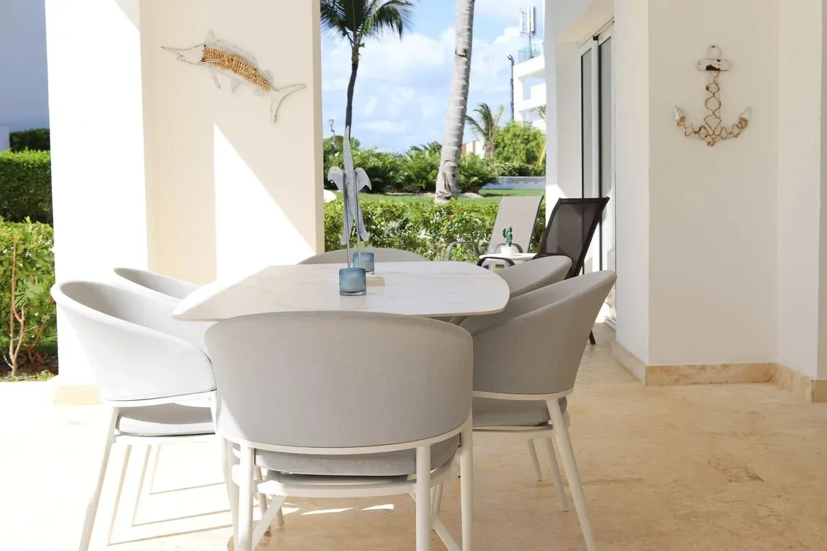 A white outdoor dining table with matching chairs sits beside a garden and palm trees.