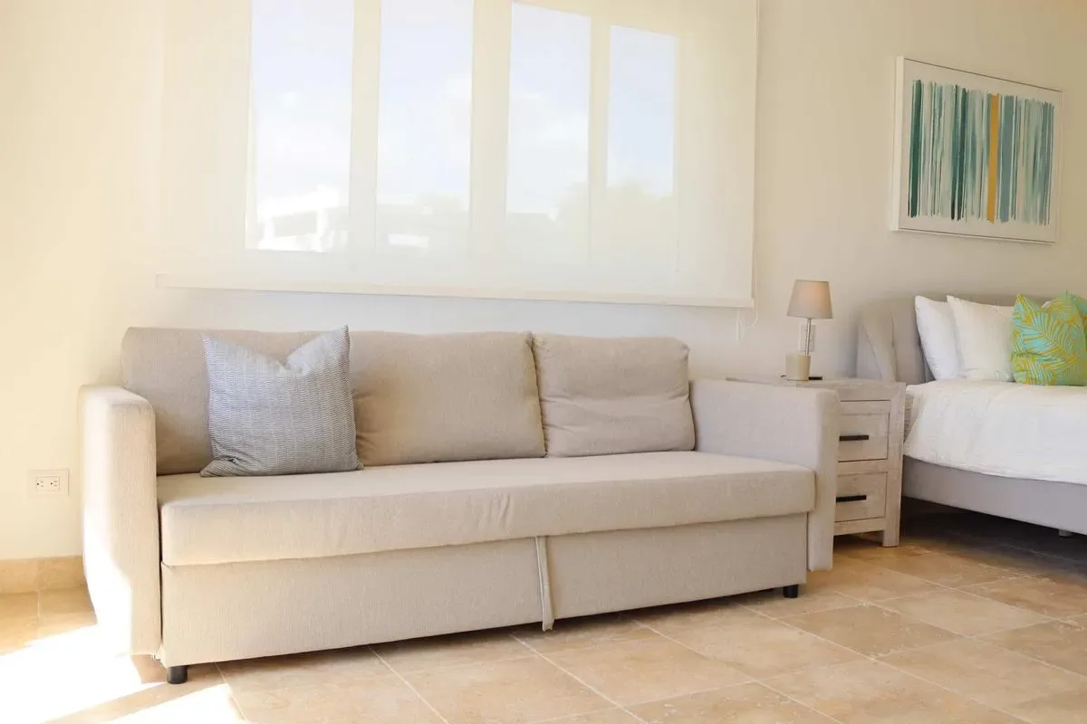 A bright bedroom has a beige sofa beneath a wide window, with a white bed and bedside table.