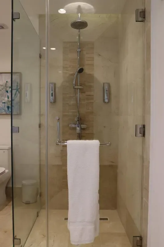 A glass-enclosed shower has a rainfall showerhead, handheld sprayer, tiled walls, and a white towel on the rail.