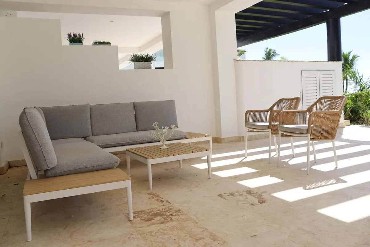 A bright covered patio has a gray sectional sofa, a low coffee table, and wicker chairs.