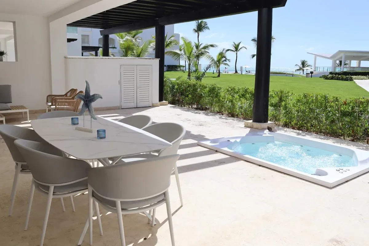 A covered outdoor dining area overlooks a small rectangular pool, manicured lawn, and palm trees.
