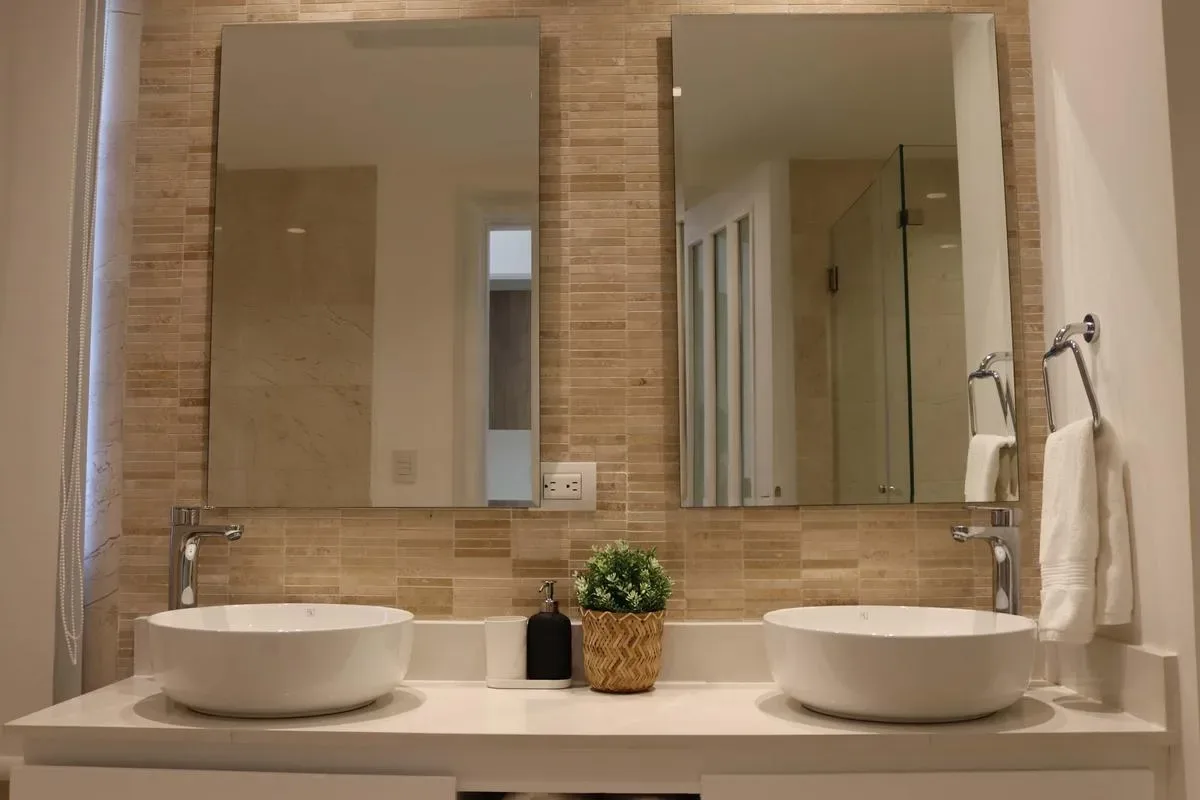 A double-sink bathroom vanity with two mirrors, tan tile walls, and a small plant between the basins.