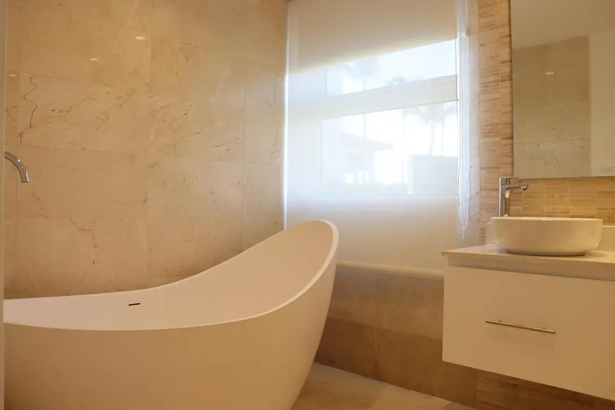 A freestanding white bathtub sits beside a bright window in a beige marble bathroom.