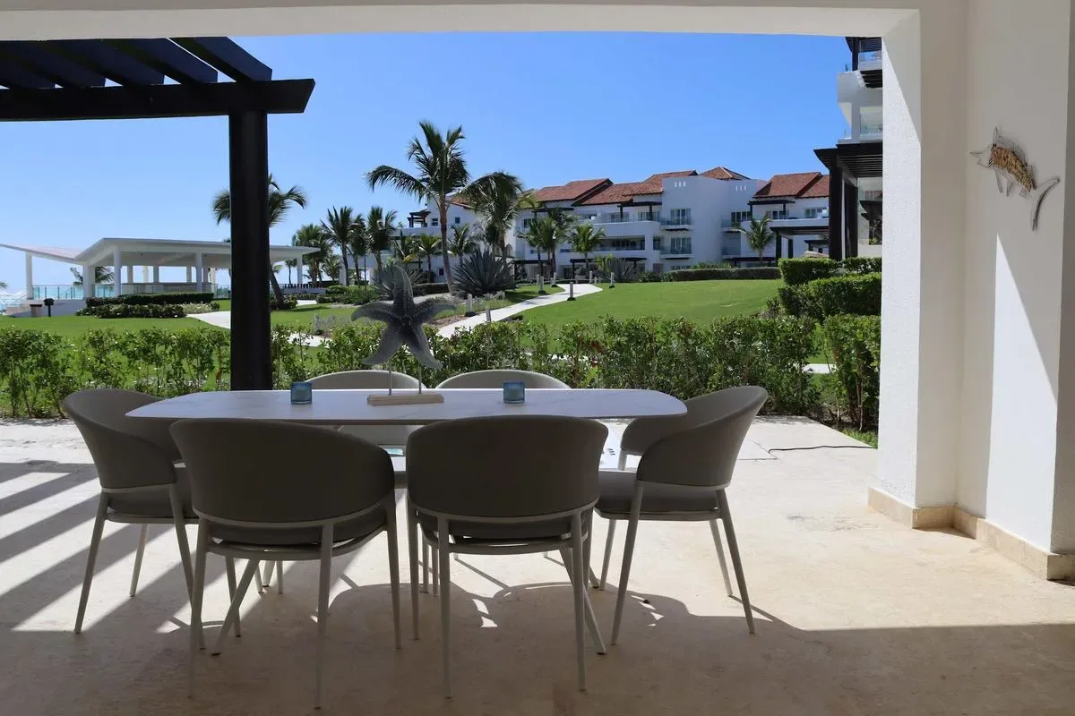 A shaded outdoor dining table with chairs overlooks landscaped lawns, palm trees, and white resort buildings.