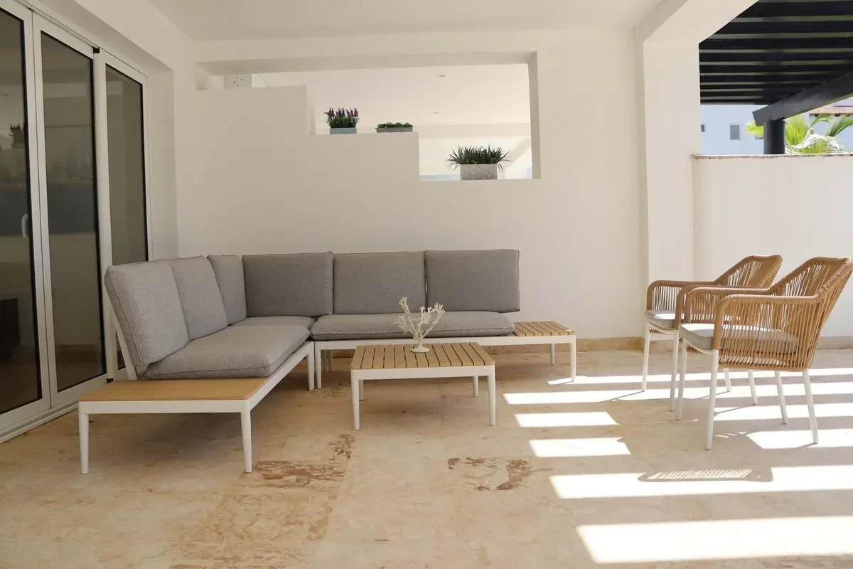 A bright covered patio with a gray sectional sofa, a low coffee table, and wicker chairs.