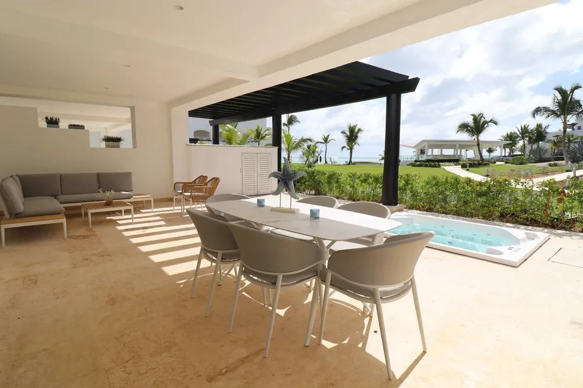 A covered patio has a dining table and lounge seating beside a small pool, with palms beyond.