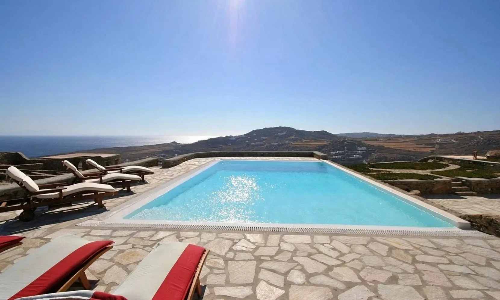 A swimming pool and surrounding sun loungers overlook a coastal landscape.